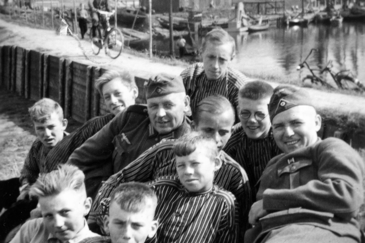 Twee Duitse militairen en acht jongens liggen lachend op de voorgrond. Op de achtergrond liggen enkele botters in de haven en komt een man met pet aanfietsen.