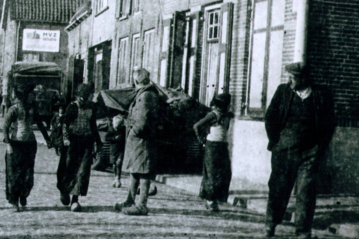 Twee mannen en drie meisjes komen aanlopen over de Oude Schans, terwijl ze achterom kijken. Op de achtergrond staat een tank tegen de huizen geparkeerd en komen nog een jongetje en meisje aanlopen.