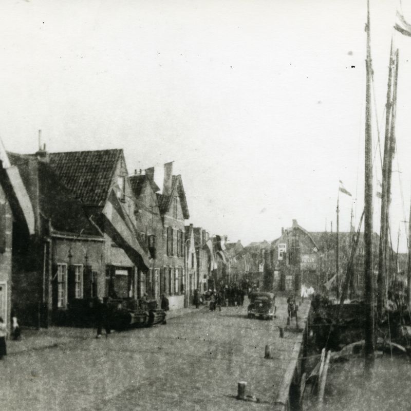 De Oude Schans met de Oude Haven in Spakenburg. Aan de huizen links en in de masten van de schepen rechts hangen Nederlandse vlaggen. Links staan voor een huis twee tanks geparkeerd, er lopen mensen op straat en er rijdt een auto.