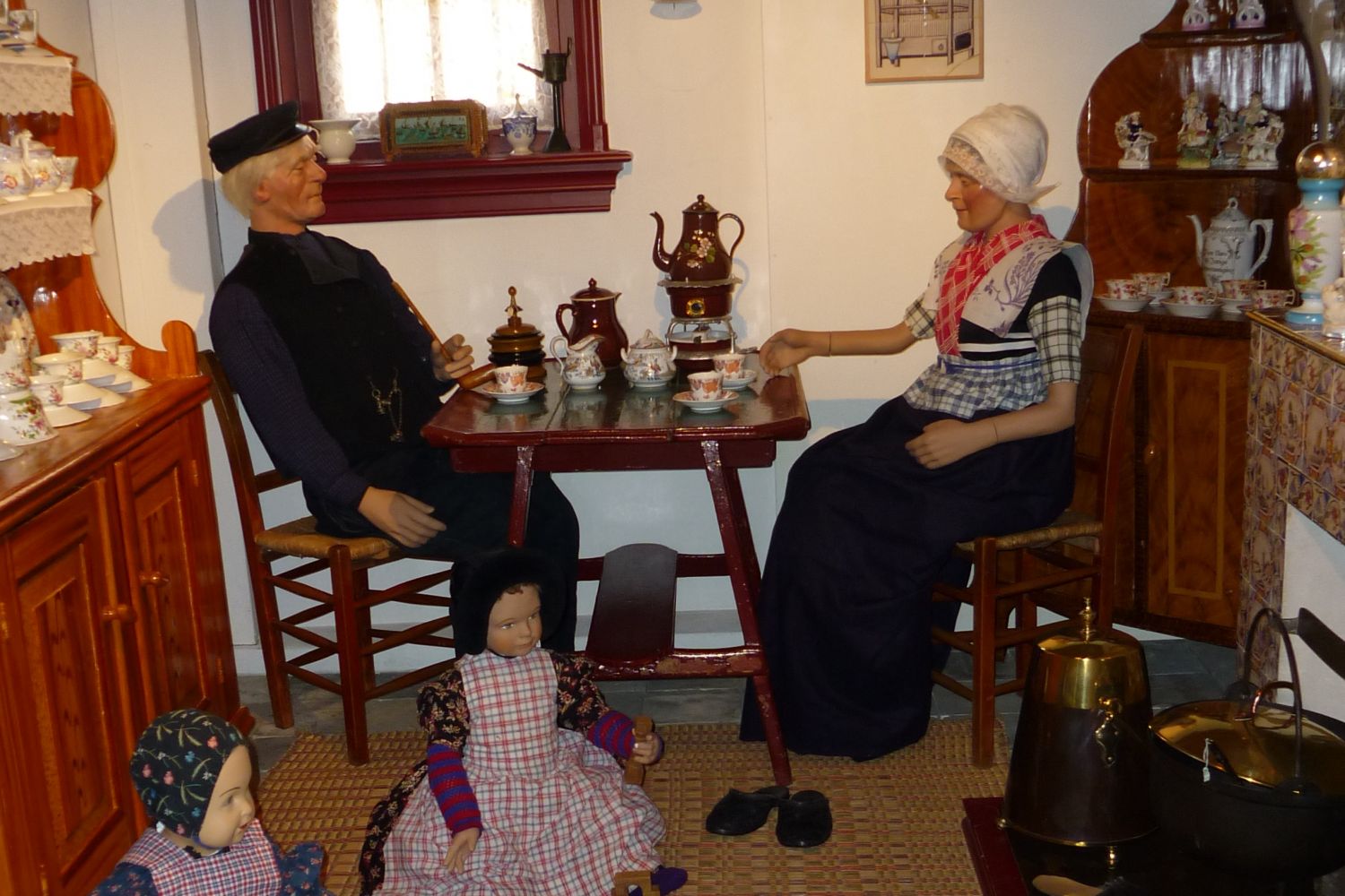 Een boer en een boerin in Bunschoter klederdracht zitten in de keuken aan tafel met een kopje koffie terwijl twee kinderen op de grond zitten te spelen.