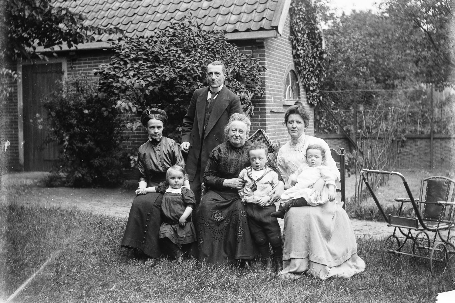 Vier volwassenen en drie kleine kinderen poseren buiten in het gras voor een schuur voor de fotograaf. De drie vrouwen zitten op een stoel. Achter hen staat een man. Op de voorgrond staan en zitten de kinderen. Rechts staat een kinderwagen.