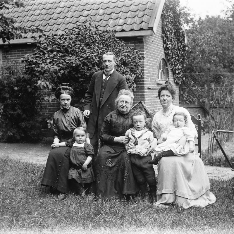 Vier volwassenen en drie kleine kinderen poseren buiten in het gras voor een schuur voor de fotograaf. De drie vrouwen zitten op een stoel. Achter hen staat een man. Op de voorgrond staan en zitten de kinderen. Rechts staat een kinderwagen.