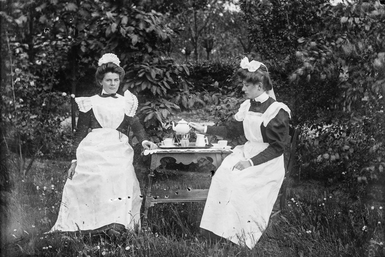 Twee vrouwen met witte schorten en witte mutsjes zitten buiten aan een tafel met een theeservies erop.