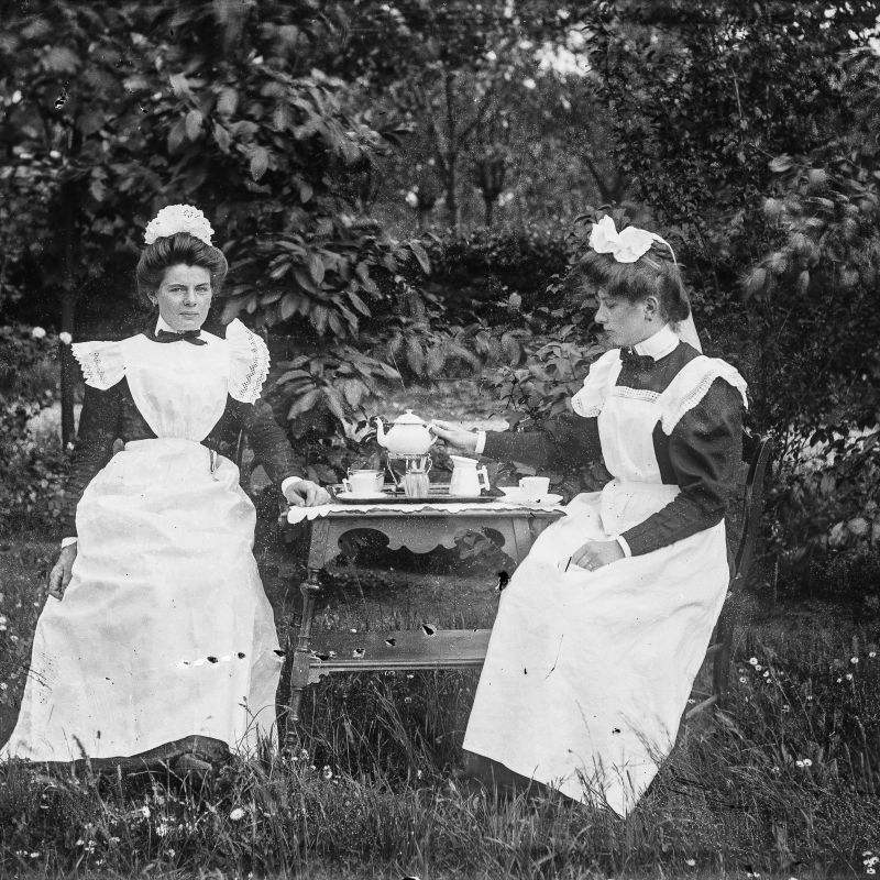 Twee vrouwen met witte schorten en witte mutsjes zitten buiten aan een tafel met een theeservies erop.
